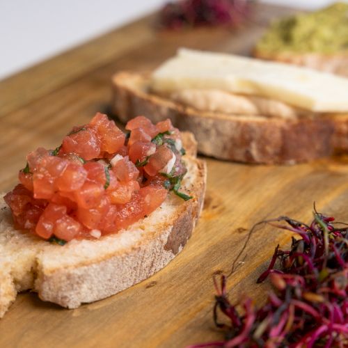 Slices of bread topped with diced tomatoes, cheese, and greens are on a wooden board, accompanied by microgreens.