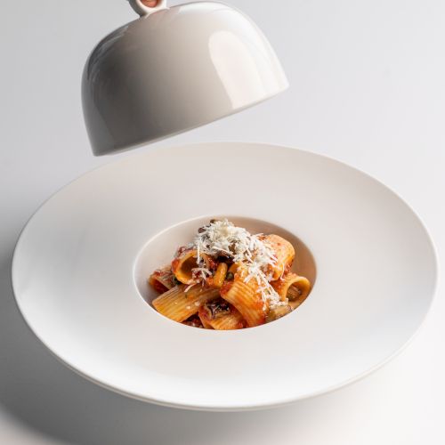 A hand lifts a white dome cover, revealing a plate of pasta topped with grated cheese in the center of a wide-rimmed white dish.
