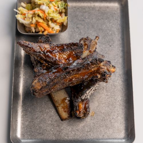 The image shows grilled ribs on a metal tray with a side of coleslaw in a small square dish.