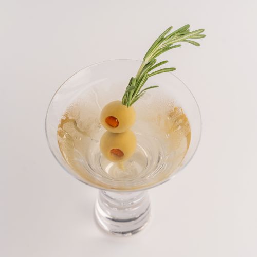 A martini glass filled with a clear cocktail, garnished with two olives and a sprig of rosemary, is shown from above.