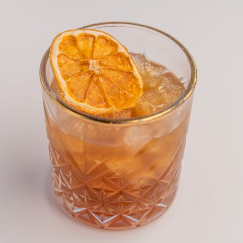 A glass of iced cocktail with a dried orange slice garnish, in a decorative tumbler, on a plain background.